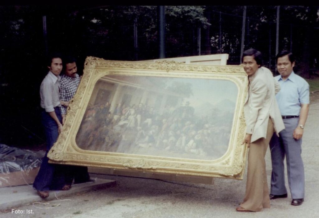 Whatsapp image 20260616 at 11 31 14 catrawarta Four men outdoors carry a large gold framed painting showing a crowd in a group photo